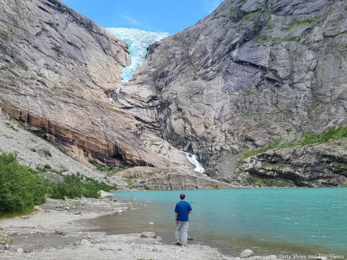 A man stands on the shore of a bright blue lake beneath a glacier at the Briksdal Glacier in Norway