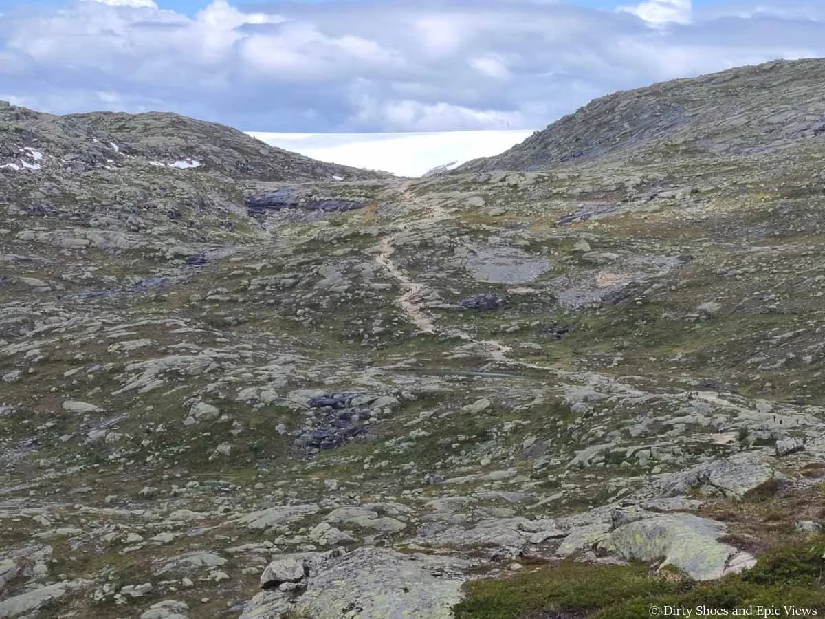 A narrow dirt path weaves its way up a rocky meadow to the top of a pass on the Trolltunga hike