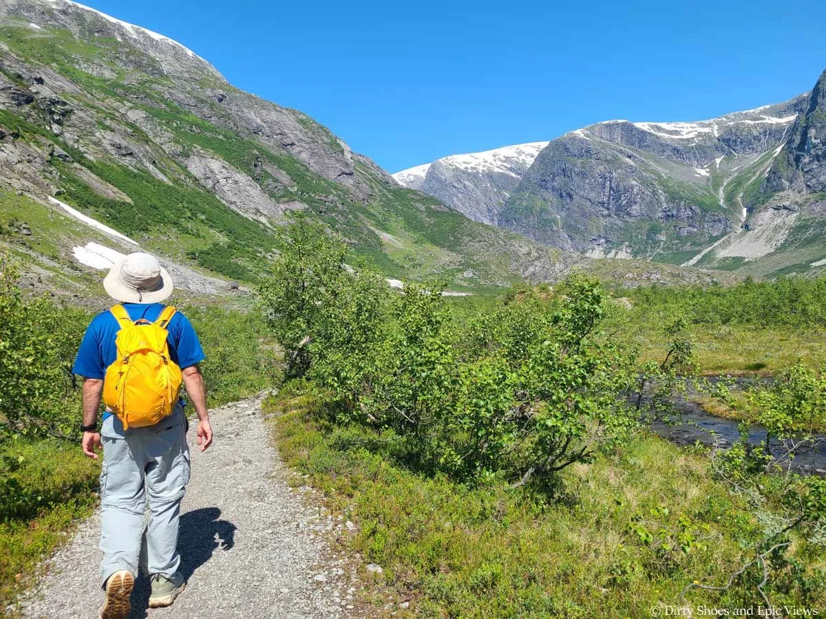 A hiker walks along a wide gravel path near a stream and mountains on the Austerdalsbreen hike