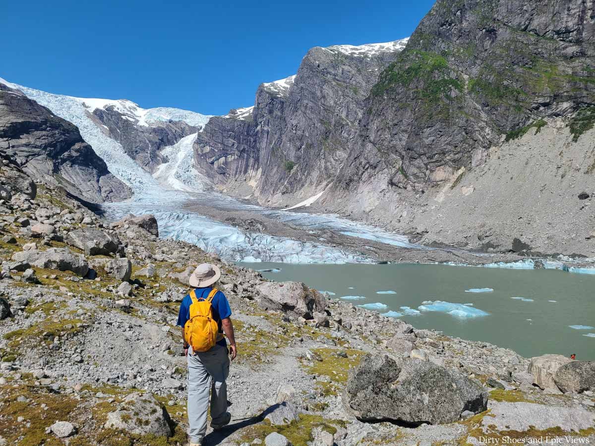 A hiker walks through rocky terrain by a glacial lagoon overlooking a large glacier arm at Austerdalsbreen in Norway