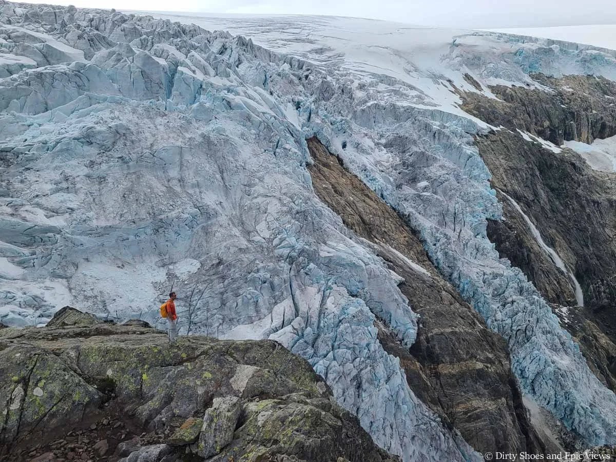 A hiker stands on a rocky outcrop overlooking a large glacier from the Reinanuten viewpoint in Norway