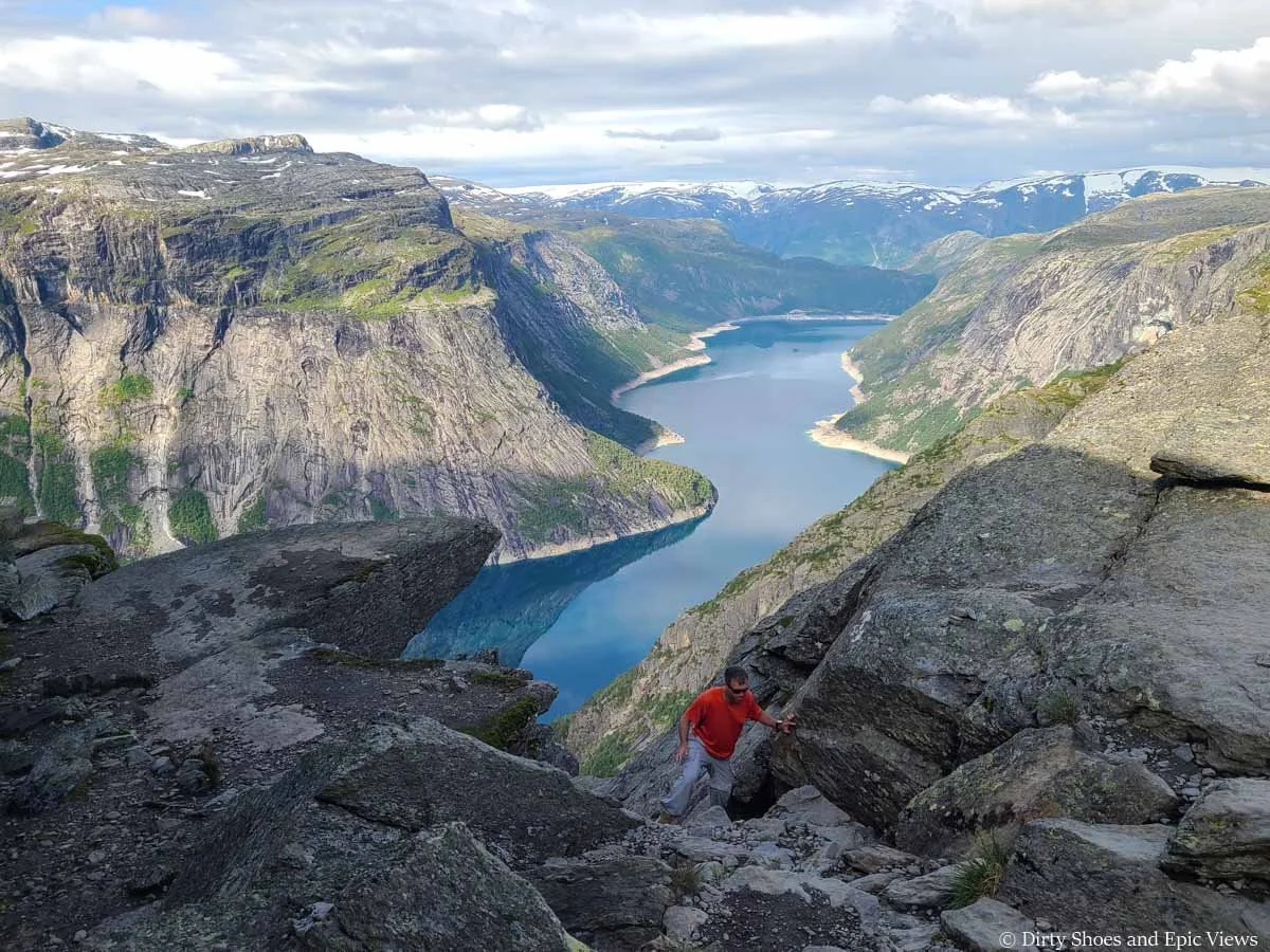 A hiker scrambles up a rocky landscape in front of lake and mountain views at Trolltunga