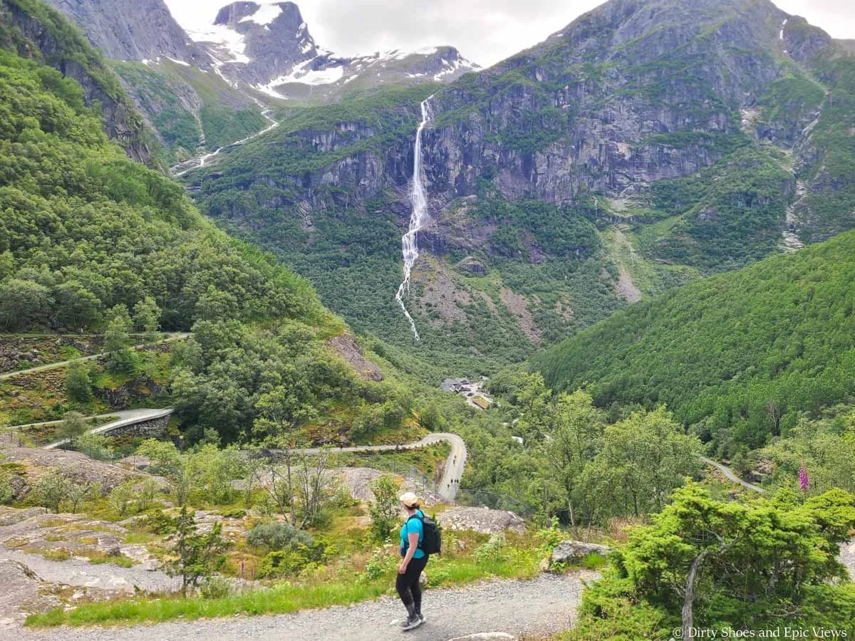 A hiker walks a path above views of a winding road and a mountain backdrop along the Briksdal Glacier trail