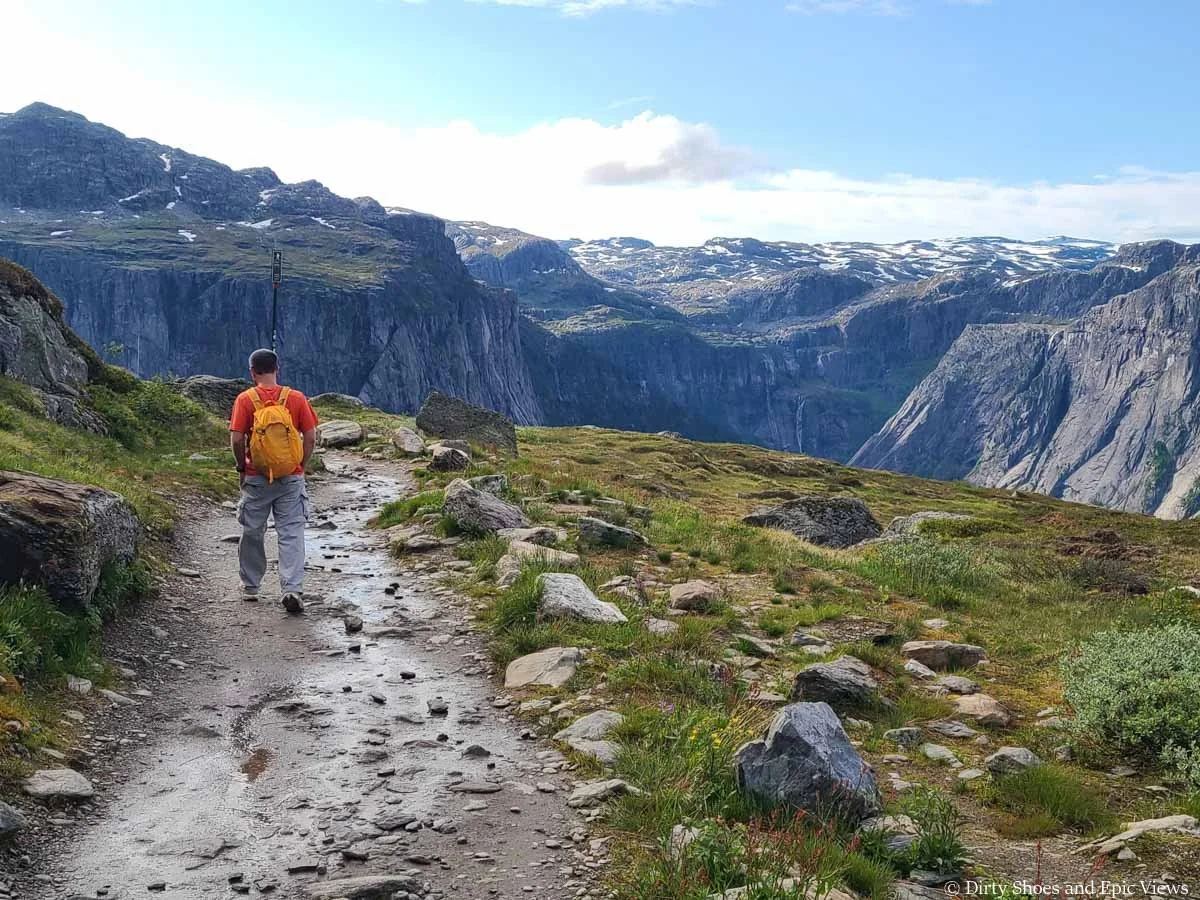 A hiker walks along a wet dirt path with views of cliffs and mountains on the Trolltunga hike