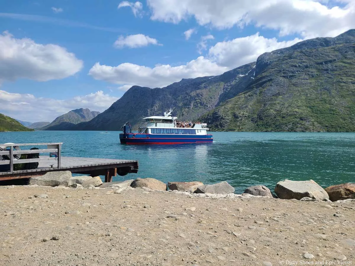 A ferry approaches a dock on a blue lake surrounded by mountains at the Besseggen Ridge trailhead