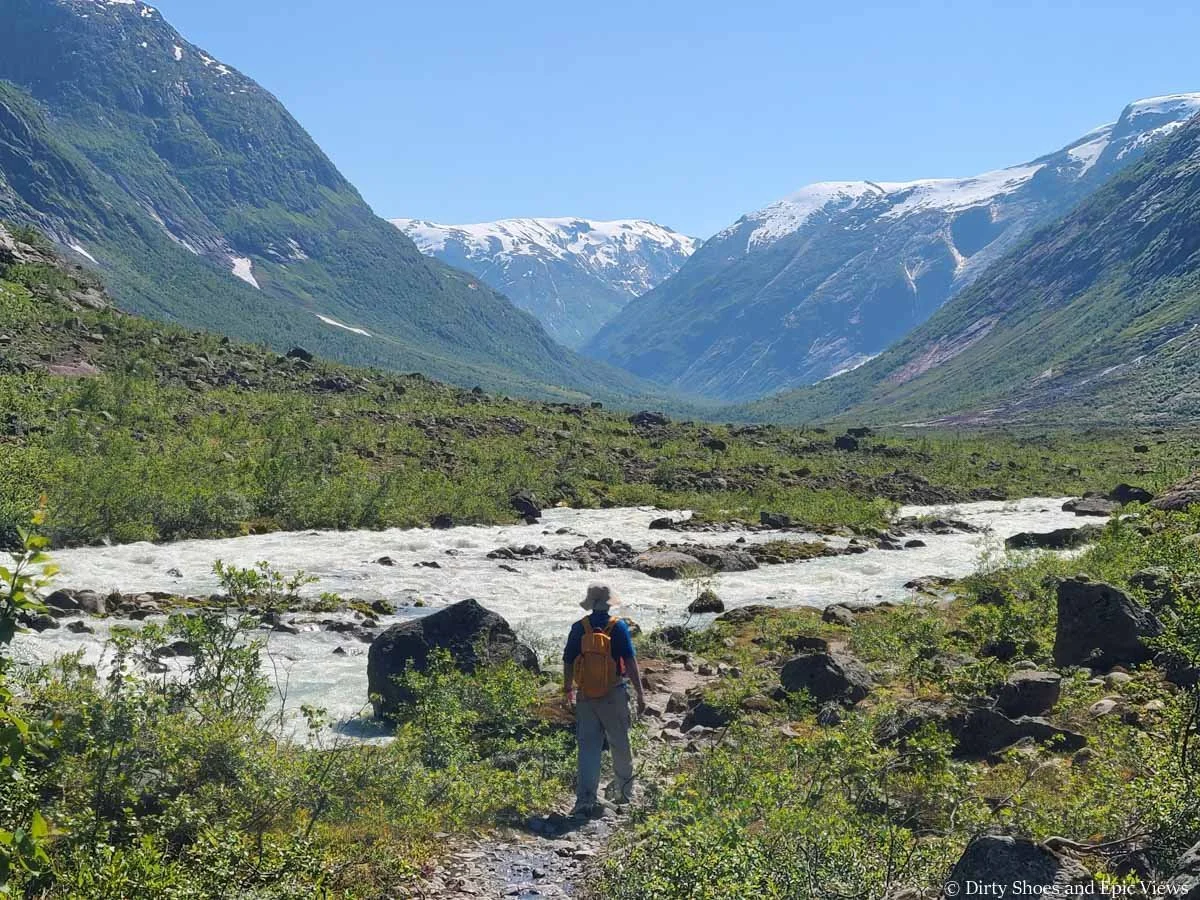 A hiker walks along a river towards snow capped mountains on the Austerdalsbreen hike