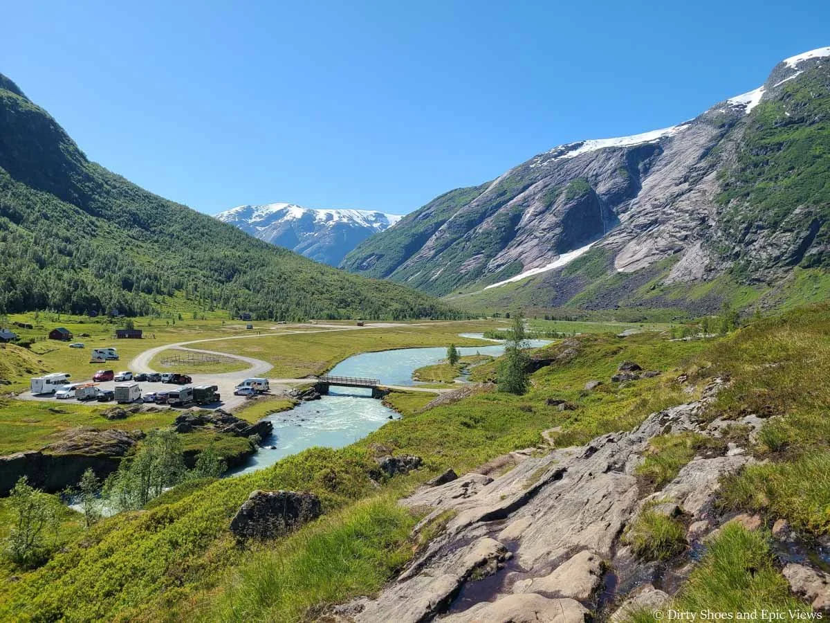 A small parking lots sits by a blue stream in a valley surrounded by mountains at the Austerdalsbreen trailhead