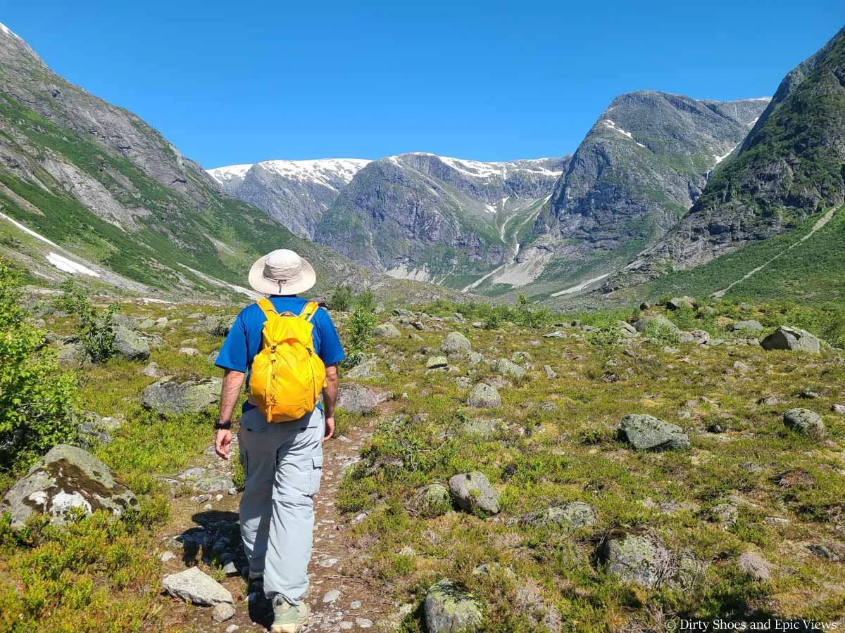 A hiker walks a narrow herd path through a rocky meadow towards mountain views on the Austerdalsbreen hike