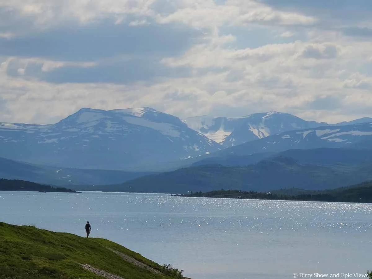 A person walks along a grassy ridge towards a lake with mountain views