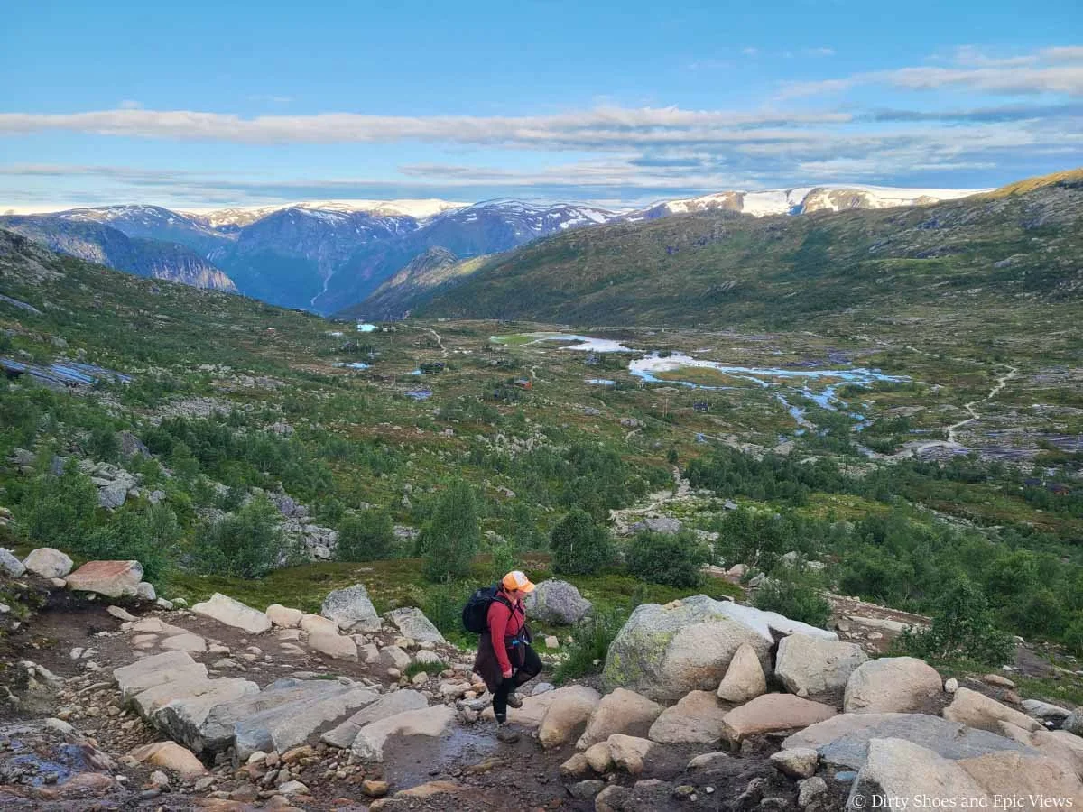 A hiker ascends steep stone steps above a grassy meadow and mountain views on the Trolltunga hike