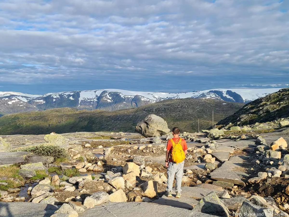 A hiker stands on a rocky path and looks out towards snowcapped mountains from the Trolltunga hike