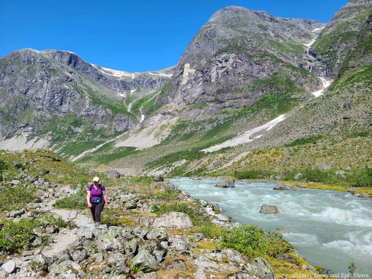 A hiker navigates a rocky landscape along a rushing blue river with mountain views on the Austerdalsbreen hike in Norway