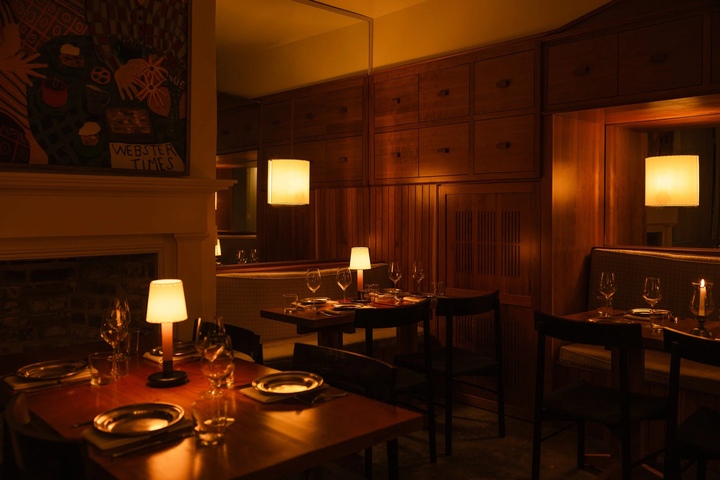 A cozy restaurant interior with warm lighting and wooden furnishings, including tables set with plates, glasses, and candles.