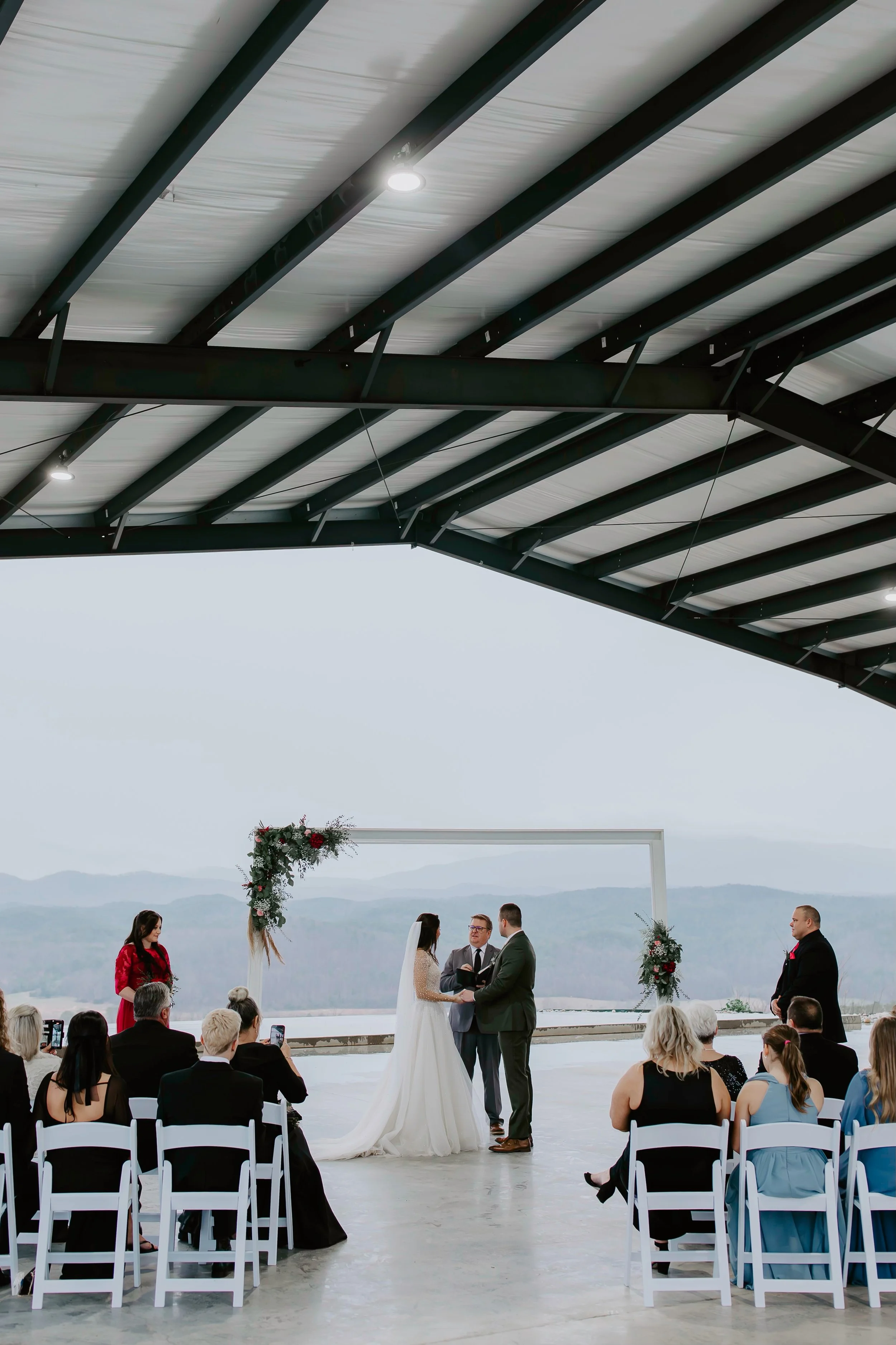 A wedding ceremony is taking place outdoors under a large, modern metal roof with a scenic mountain view in the background. The bride and groom are standing before an officiant, holding hands, with guests sitting in chairs watching. The area is decorated with floral arrangements and a white arch.