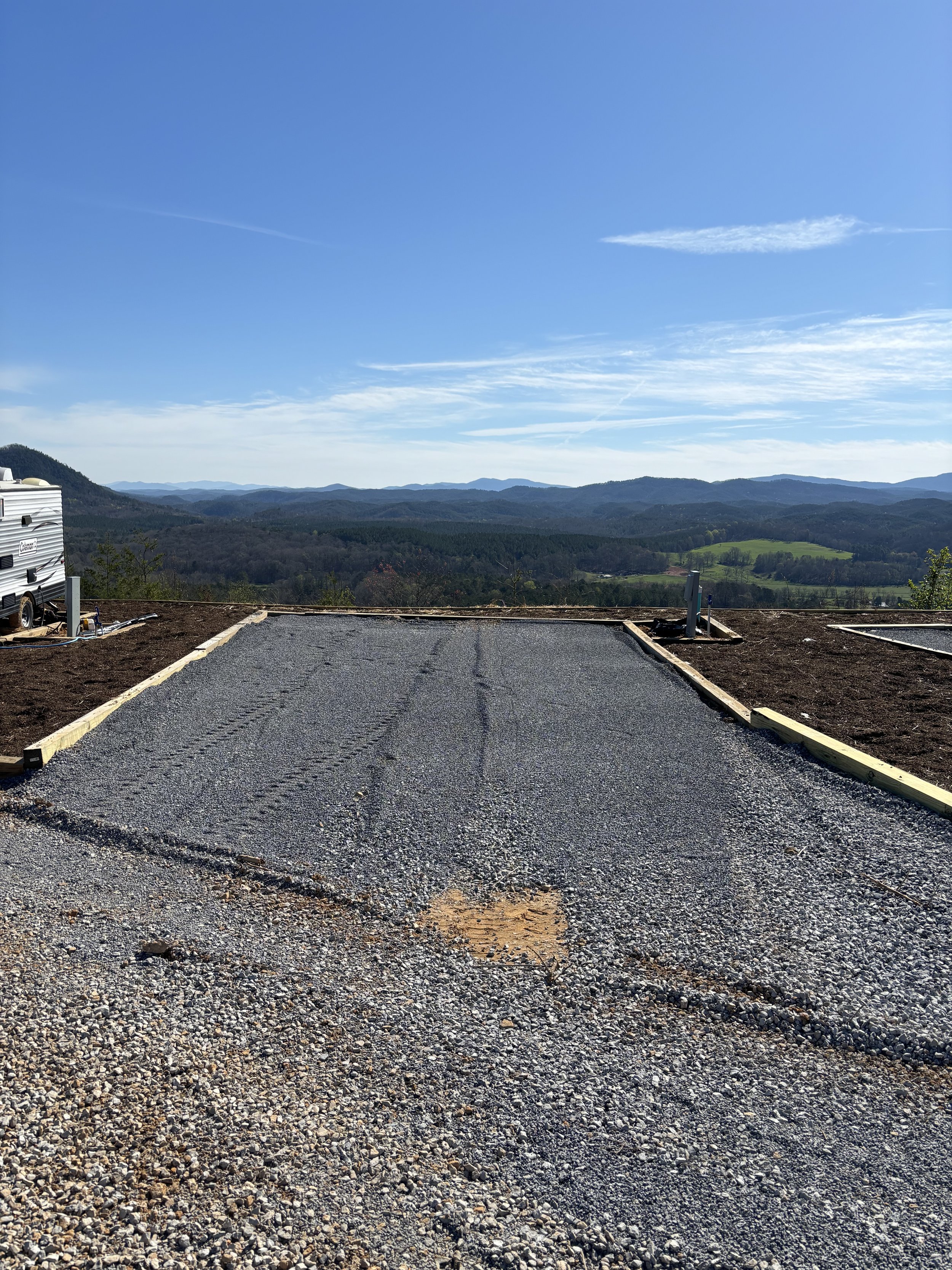 An RV site with newly poured asphalt and wooden borders, overlooking a scenic mountain landscape with blue sky and clouds.
