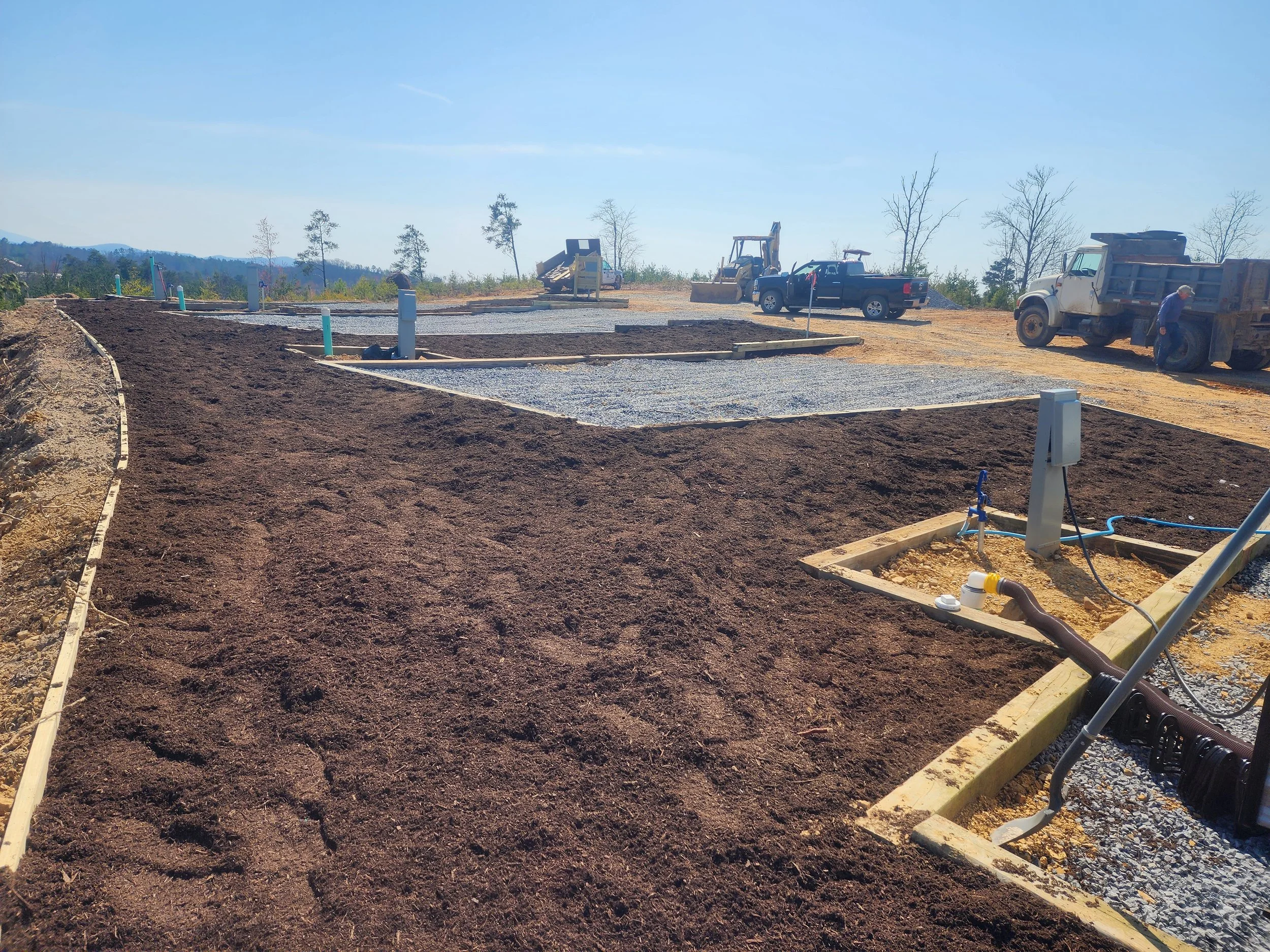 An RV site with dirt and gravel, wooden borders, pipes, a dump truck, a black pickup truck, a yellow backhoe, and a worker, set against a background of trees and clear blue sky.