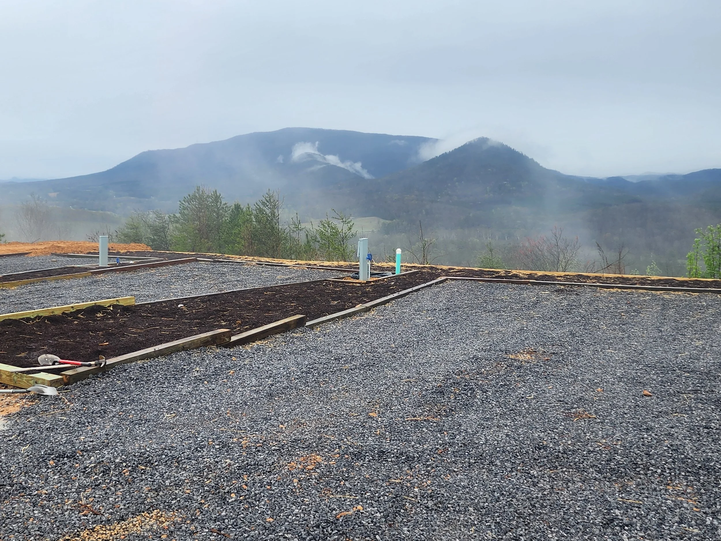An RV site with a View of mountains with some low clouds and smoke, with a foreground of ground with gravel and wooden boards, possibly a construction site.
