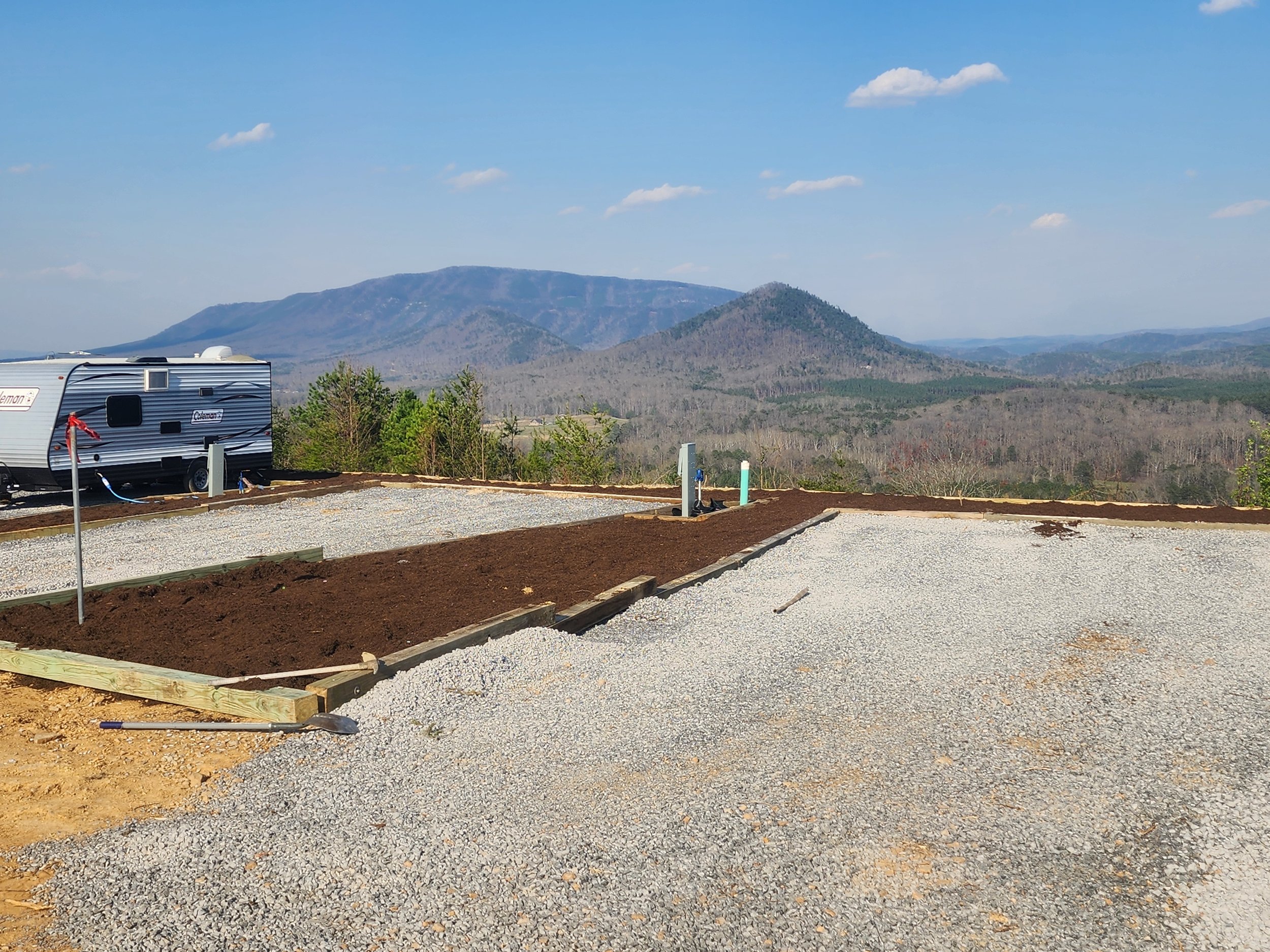 An RV site with gravel and dirt, wooden framing for a structure, and a scenic mountain landscape in the background.