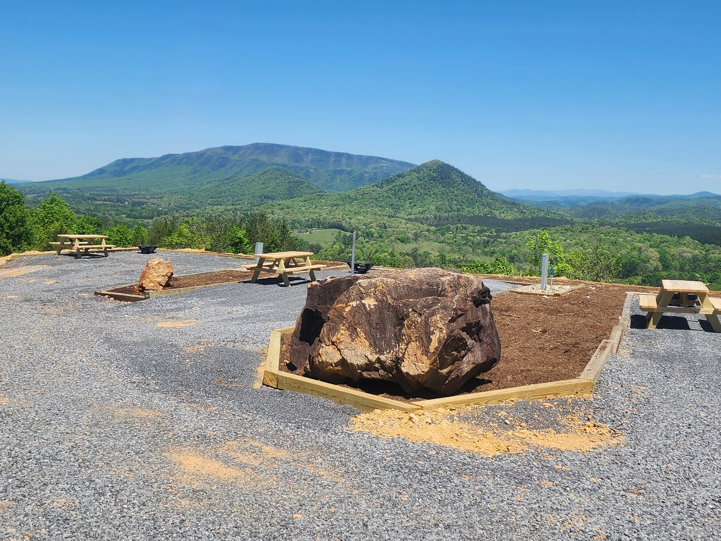 An RV site with a Scenic view of green, forested mountains with a clear blue sky, some picnic tables and large rocks in the foreground.