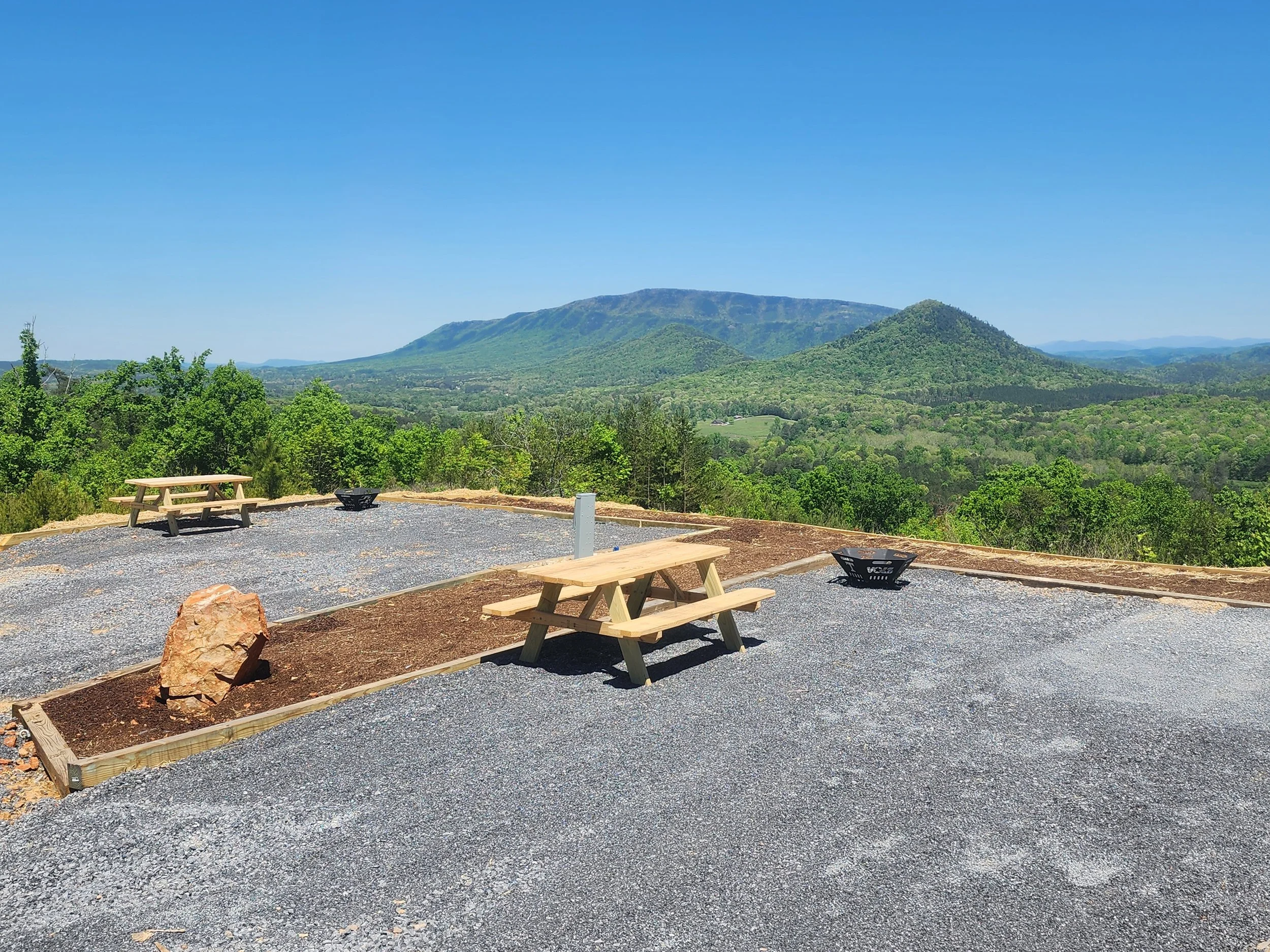 An RV site with scenic view of green mountains and lush forests under a clear blue sky, with a picnic area in the foreground featuring wooden benches, gravel ground, and small fire pits.