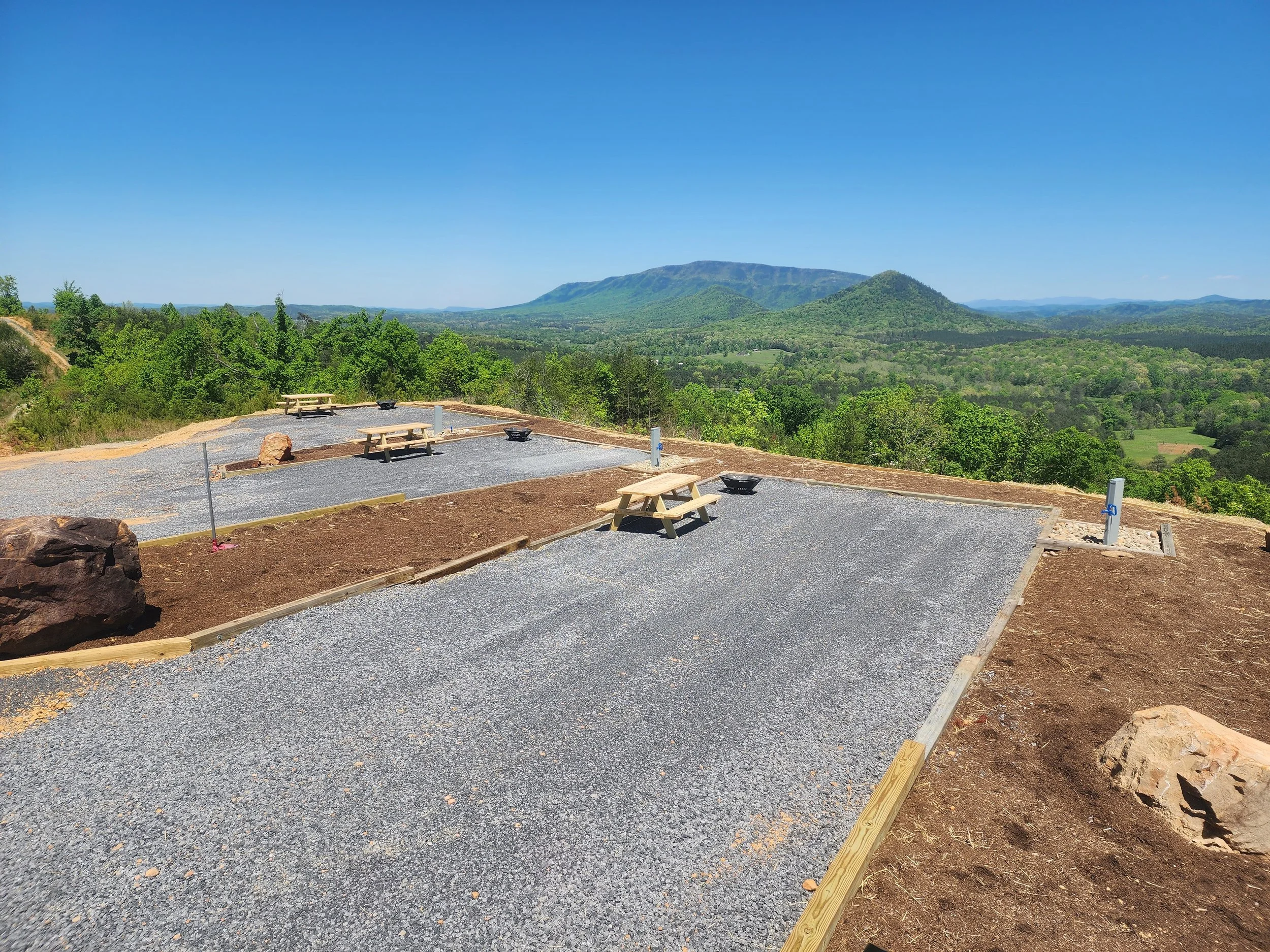 An RV site with Scenic view of green mountains under a clear blue sky with a viewing area in the foreground featuring benches and gravel pathway.