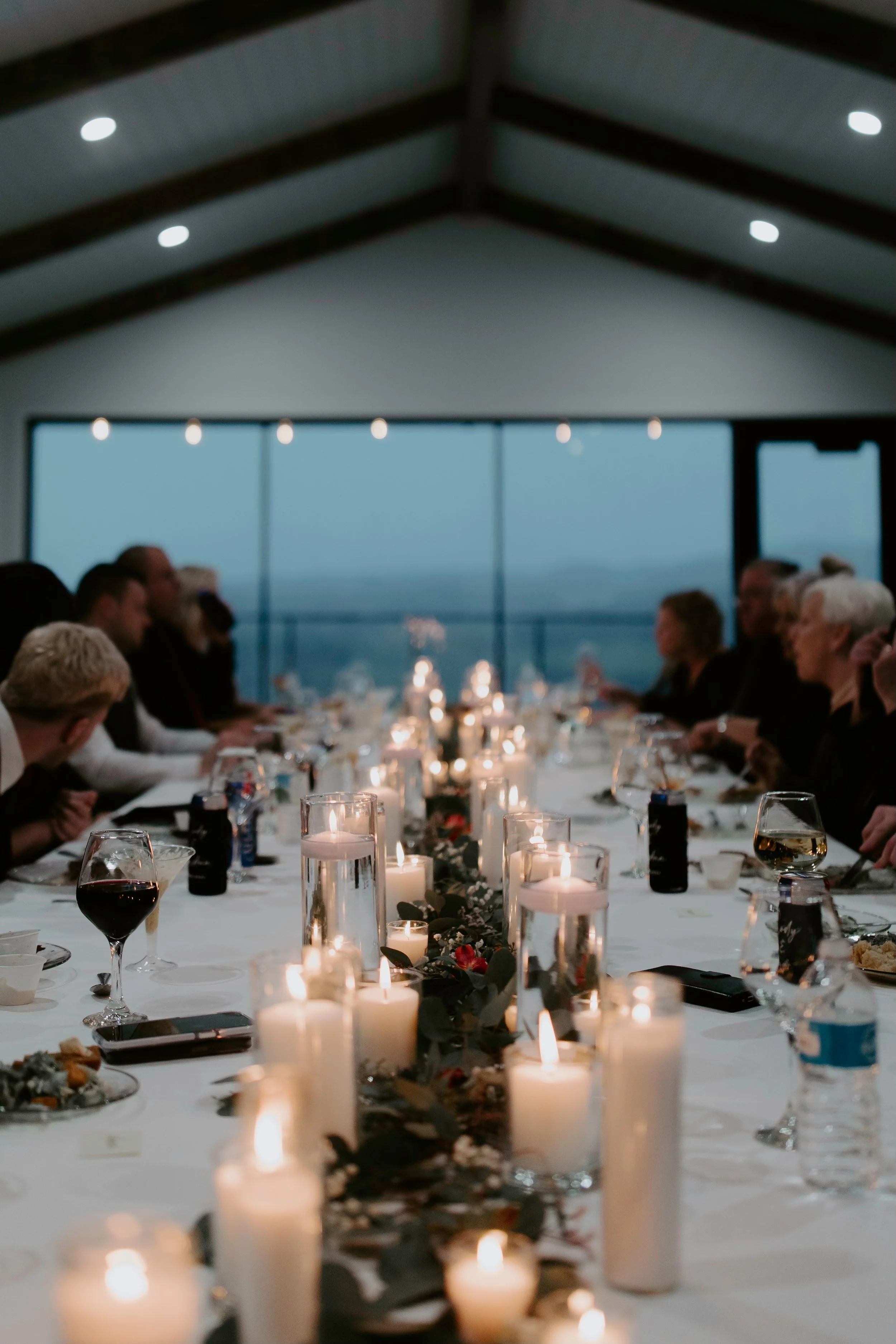 A formal dinner event inside a modern building with large windows and view of the ocean. Guests are seated around a long table decorated with white candles and greenery, enjoying drinks and conversation.