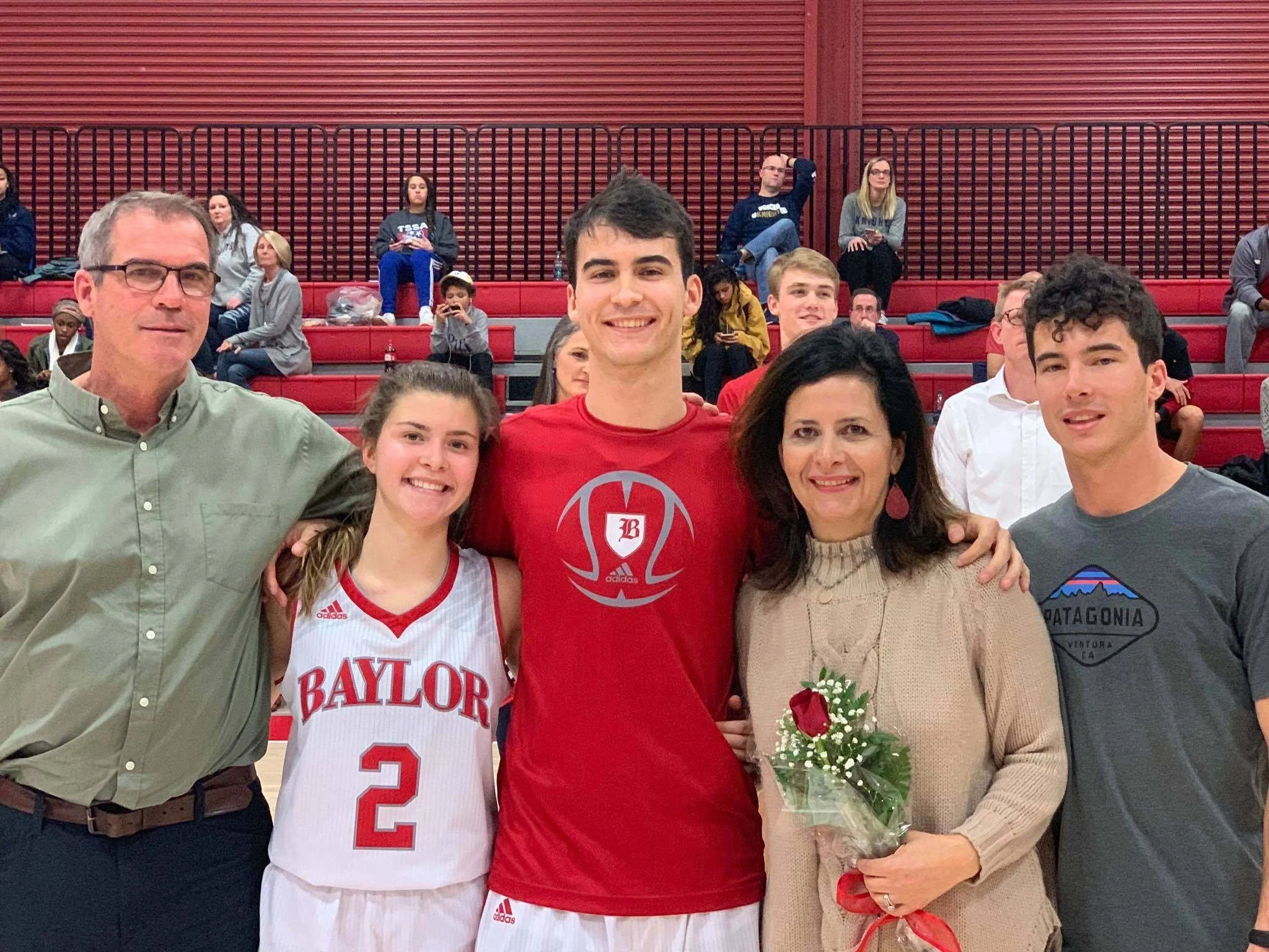 Group of smiling people at an indoor event, with a man in a gray shirt and glasses, a young woman in a basketball jersey, a woman holding flowers, and others in the background seated on red bleachers.