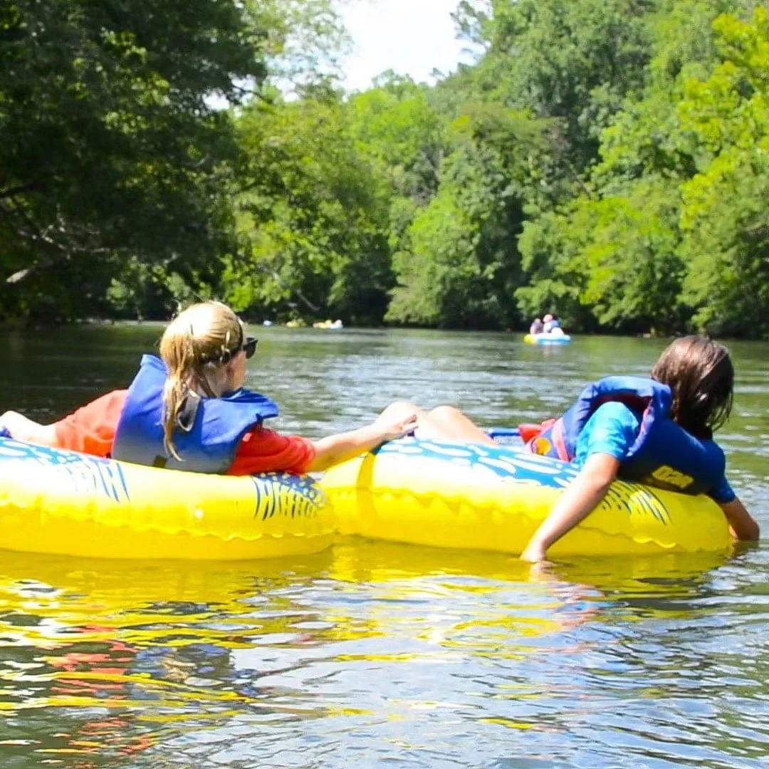 Two children wearing life jackets on a yellow inflatable tube floating on the lower ocoee river surrounded by green trees in a sunny outdoor setting.