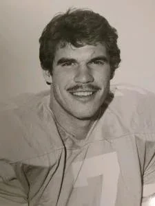 Black-and-white photo of a young man with curly hair and a mustache, smiling at the camera.