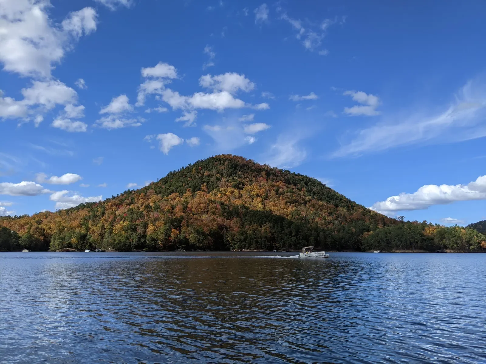 Parksville lake with a boat passing by, surrounded by a mountain covered in trees with autumn foliage, under a partly cloudy sky.