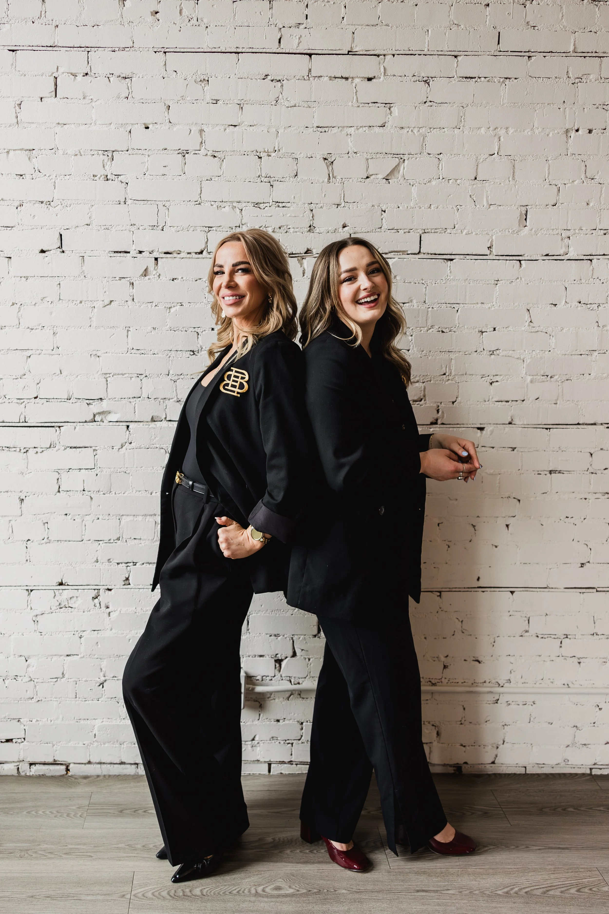 Brittany and team member, Indy, standing back to back in black blazers against a white brick wall at Beauty by B in Calgary.