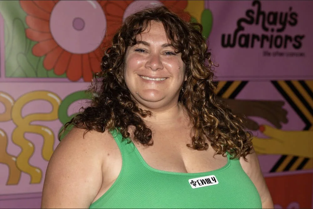 Close-up of a woman with curly brown hair, wearing a green top and a name tag that reads 'Emily', smiling in front of colorful, fun background with large floral and geometric designs.