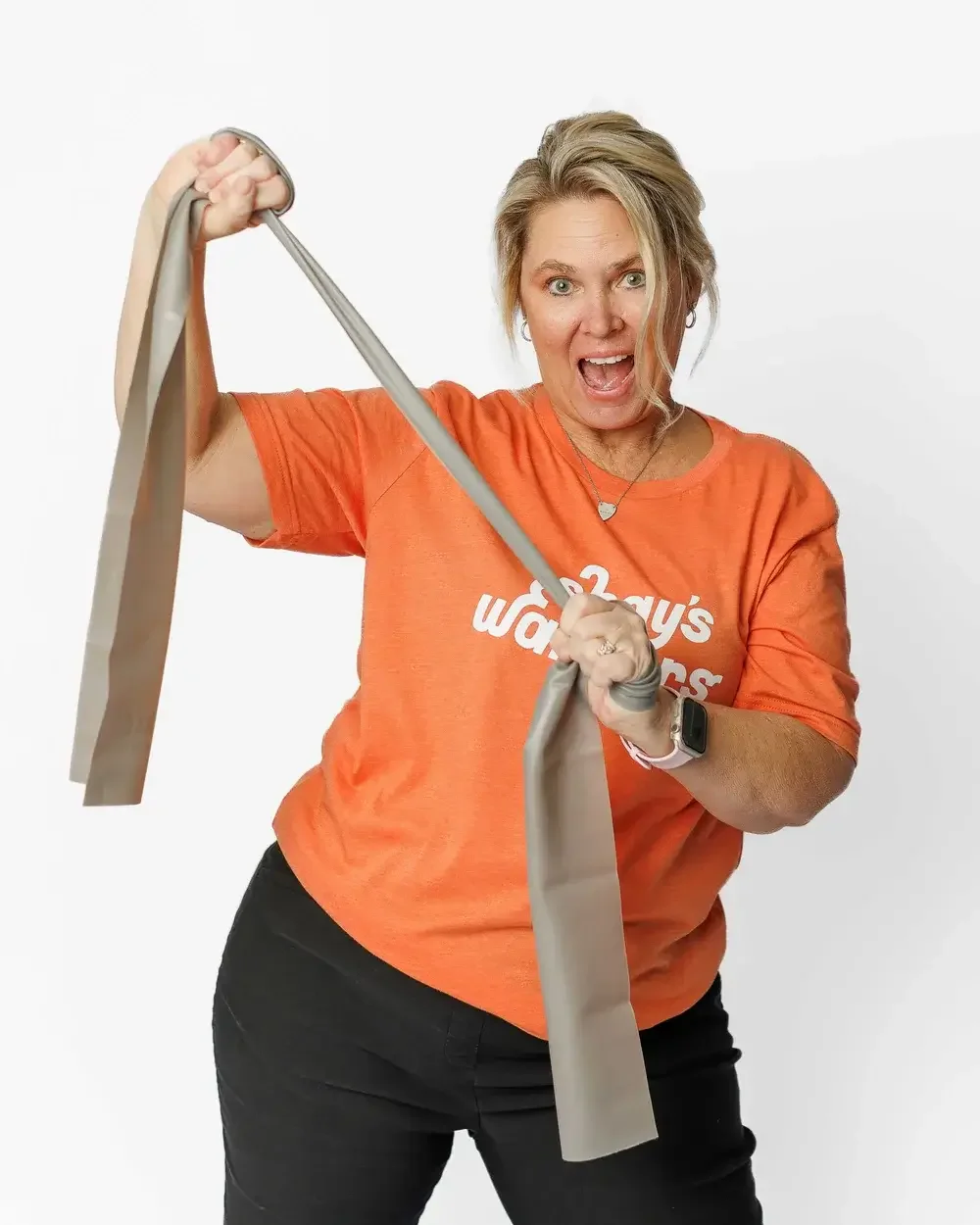 Woman in orange T-shirt that says 'Shay's Warriors life after cancer' holding a long gray strap or scarf, with an excited facial expression.