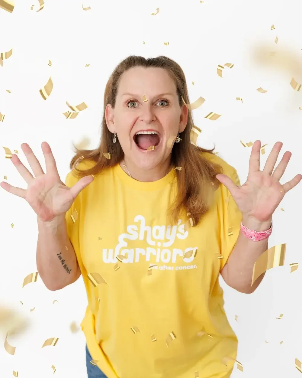 A woman wearing a yellow shirt with white text, celebrating with confetti falling around her, with her mouth open in excitement and raised hands.