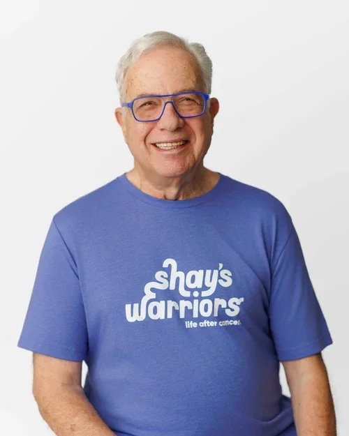 Man with short white hair wearing a blue Shay's Warriors tshirt and matching blue eyeglasses smiling at the camera