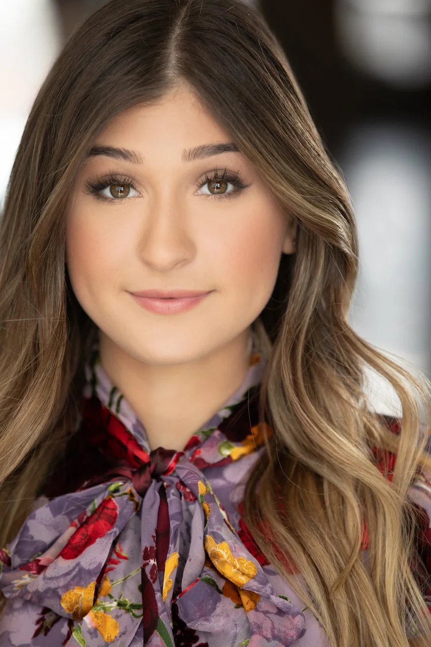 Close-up of a young woman with long wavy brown hair, wearing a floral scarf tied at the neck, smiling softly, indoors with a blurred background.