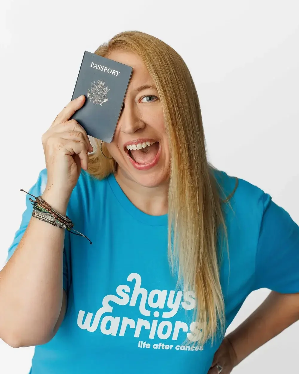 A cheerful woman with long blonde hair, wearing a blue t-shirt that says 'shays warriors life after cancer,' holds a U.S. passport up to her face, smiling widely.