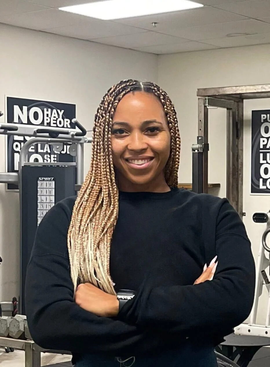 Woman with braids smiling standing with arms crossed in a gym, surrounded by exercise equipment.