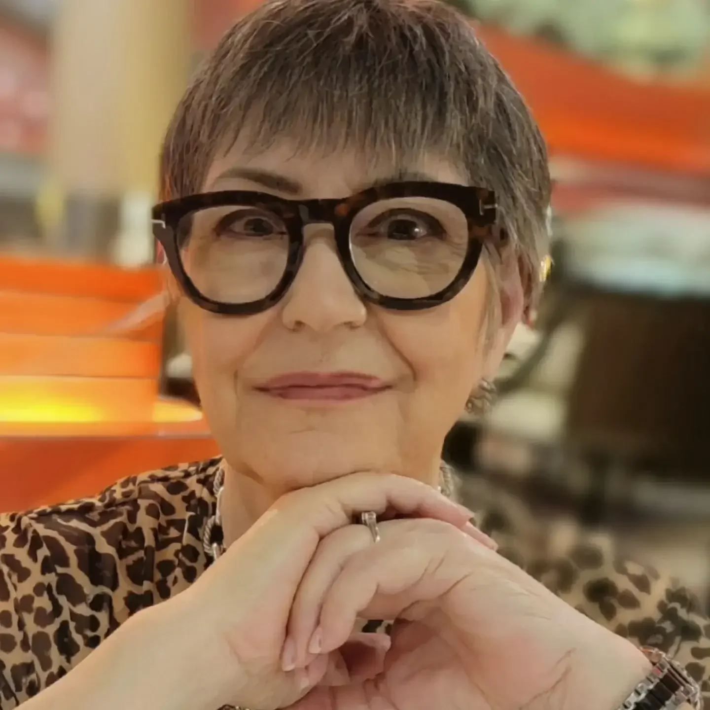 A smiling woman wearing black glasses and a leopard print blouse with her chin resting on her overlapping hands. The background behind her is blurred.