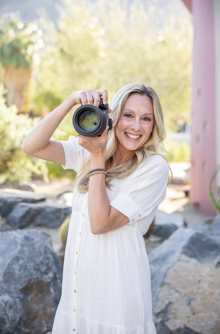 A woman with long blonde hair wearing a white dress, smiling, and holding a camera up to her face outdoors among rocks and trees.