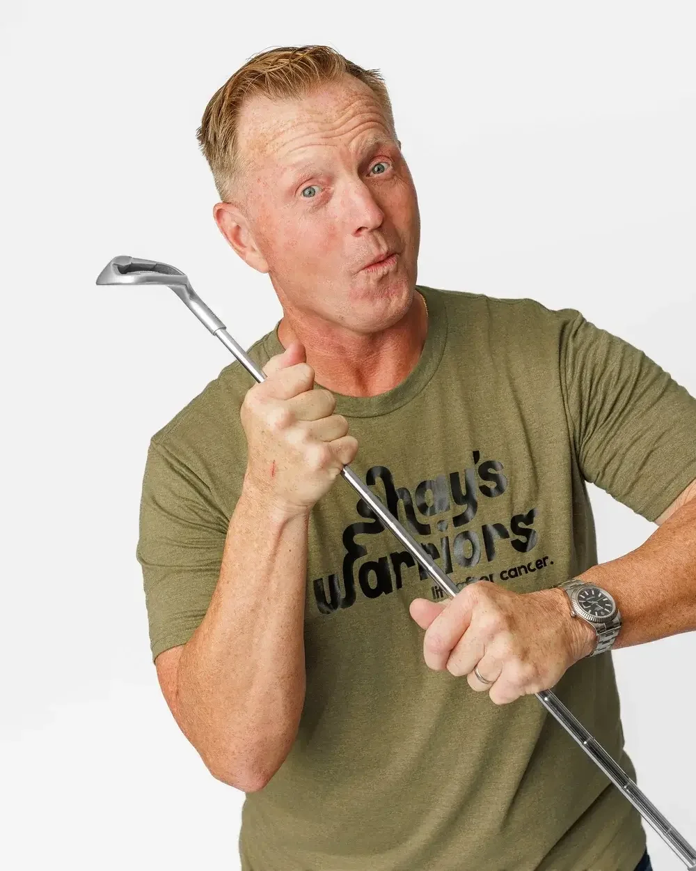 A man with blonde hair and blue eyes holding a golf club, wearing a green T-shirt with black printed text that says 'Shay's Warriors life after cancer', and a silver wristwatch, against a plain white background.