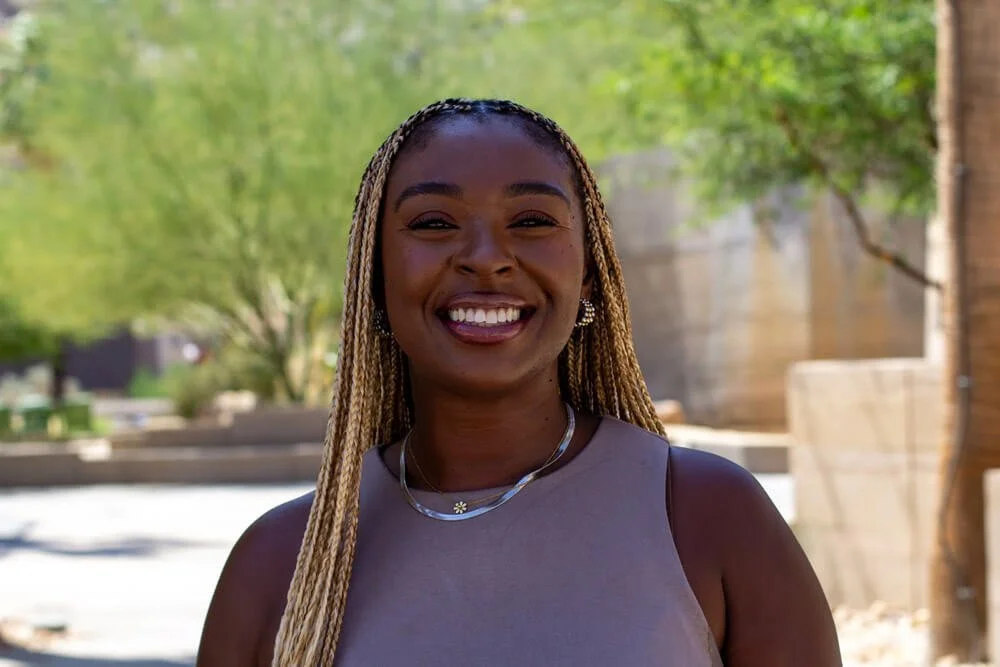 A young woman with smiling face, long blonde braided hair, wearing earrings, a necklace, and a sleeveless top, standing outdoors with blurred green trees and sunlight in the background.