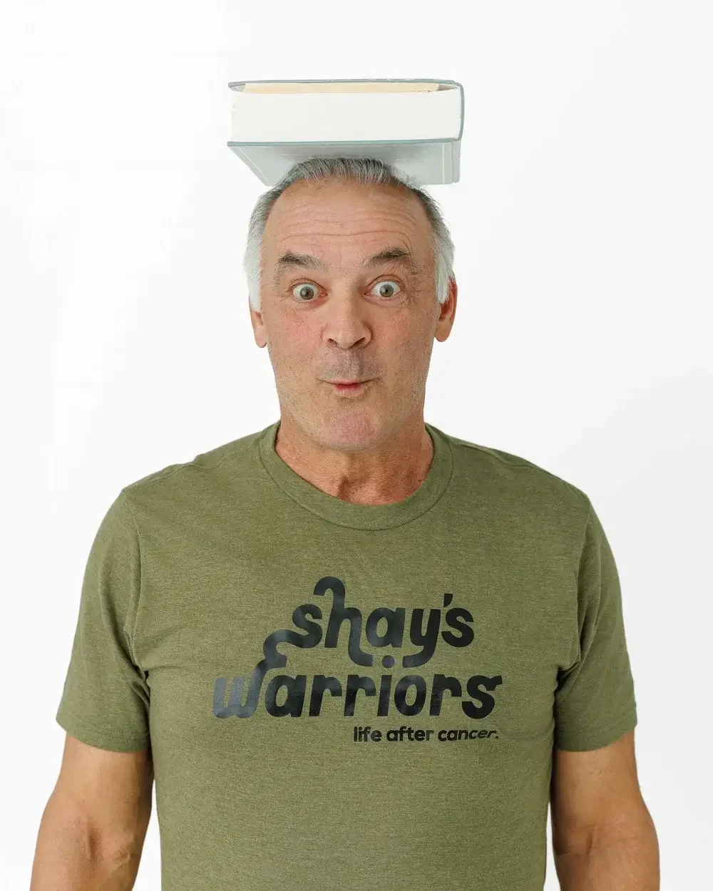 A man with gray hair and blue eyes balancing a large hardcover book on his head, standing against a plain white background.