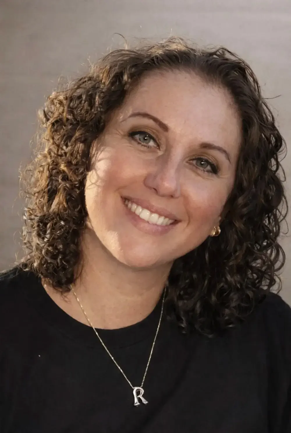 Close-up of a smiling woman with curly brown hair, light-colored eyes, wearing a black top, a gold necklace with a letter R pendant, and gold earrings.