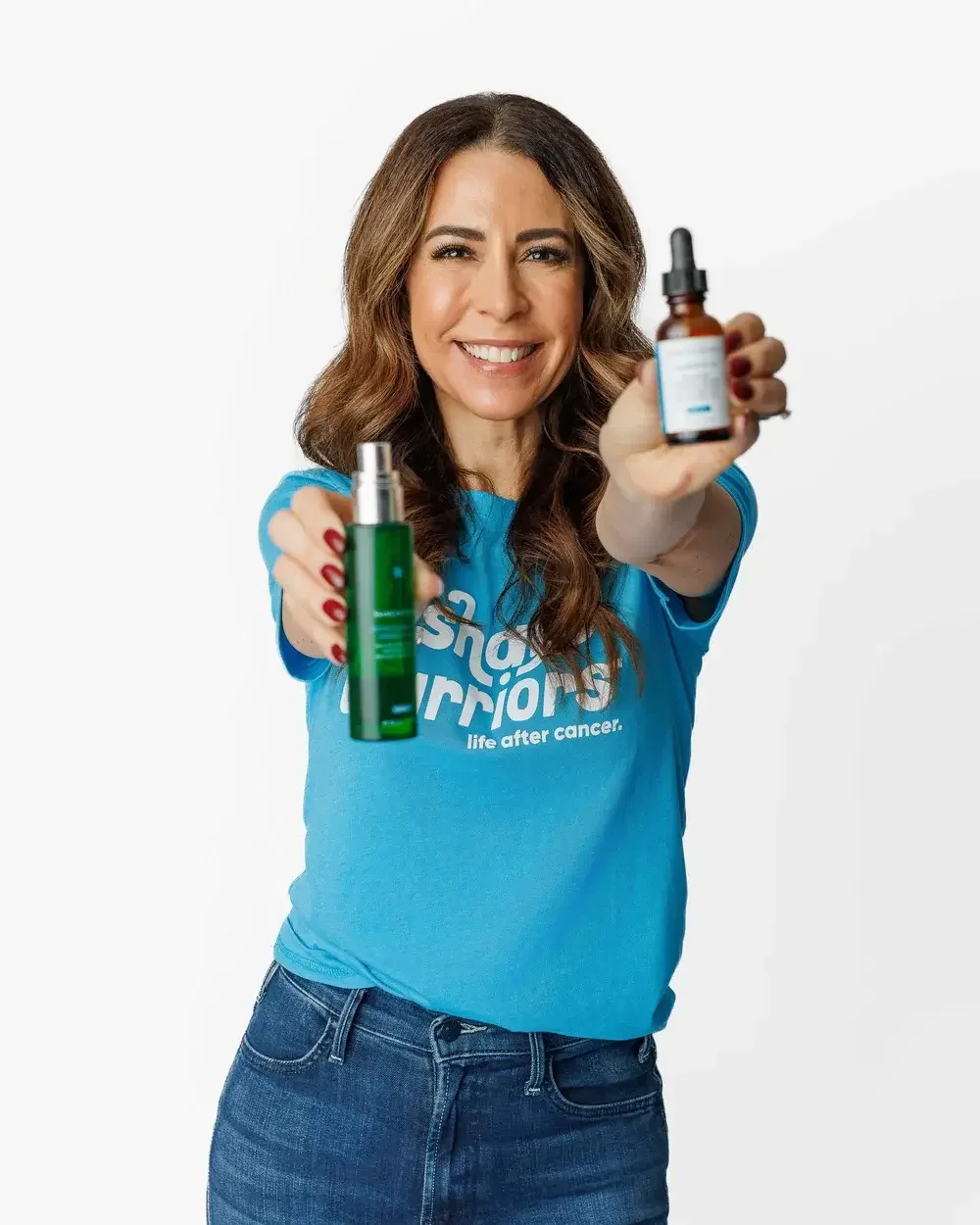 A woman smiling, wearing a blue T-shirt with white text, holding a green skincare spray bottle in one hand and a brown dropper bottle in the other, against a plain white background.