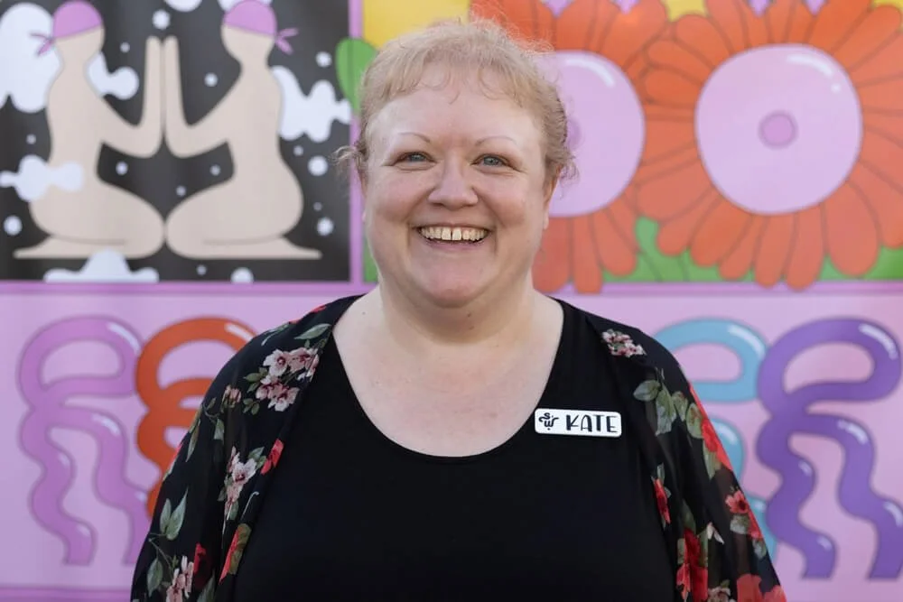 A woman with light skin and blonde hair smiling in front of colorful, abstract art background. She is wearing a black top with a floral cover-up and has a name tag that says 'KATE'.