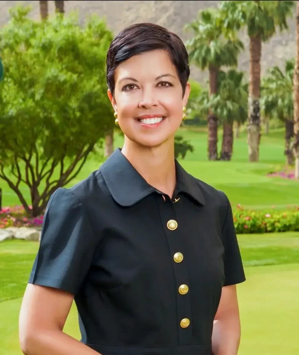 A woman with short dark hair standing outdoors in a park with trees and green grass, smiling, wearing a black short-sleeved dress with gold buttons and earrings.