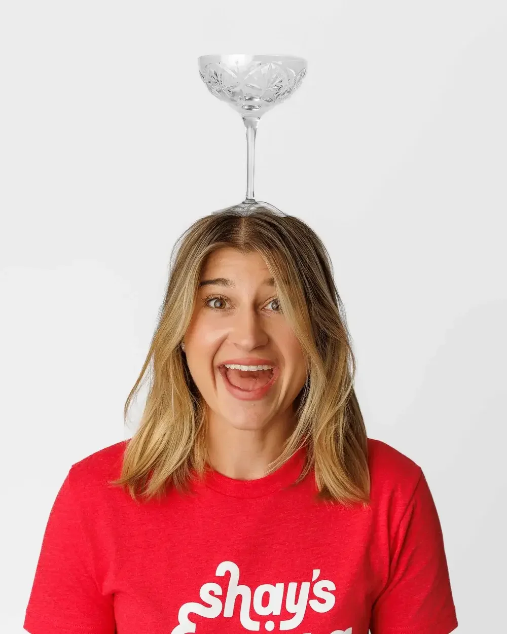 A woman with shoulder length hair balancing a martini glass on her head while wearing a red T-shirt that says 'Shay's Warriors life after cancer'.