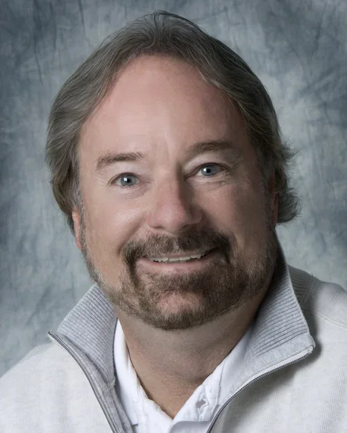 Man with grayish short hair, mustache, and beard wearing a half-zip light gray sweatshirt