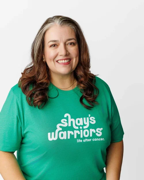 Woman with long wavy red hair with a white streak in the front wearing a green Shay's Warriors tshirt